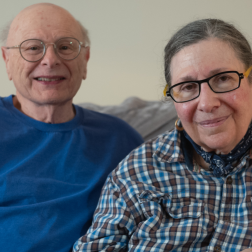 Caryn and Michael, an older Caucasian couple. He is bald with glasses and a blue shirt. She has long grey hair, glasses, and a checkered shirt. 