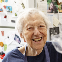 Citymeals on Wheels recipient Joyce, a senior woman with white hair. She is smiling and wearing a blue shirt in her NYC apartment. 