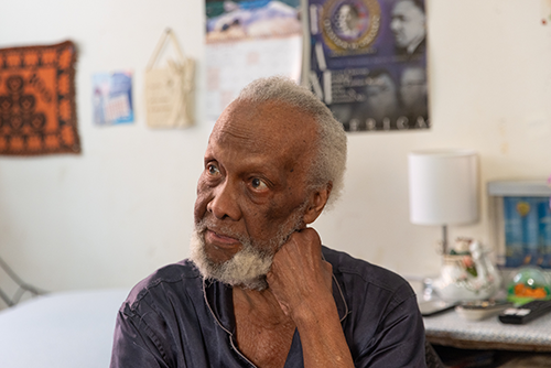 Citymeals recipient Bryan, an older man with dark skin and a long white beard wearing blue pajamas looks thoughtfully to the left of the frame. 