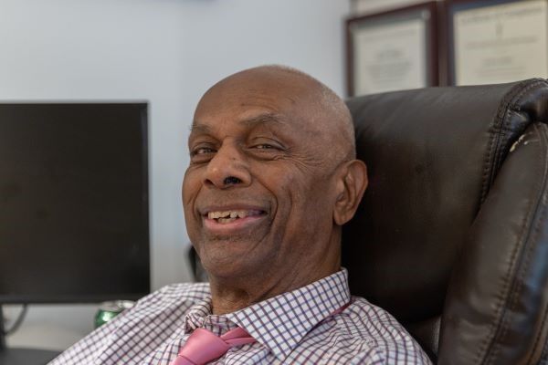 An older black man sits in an office chair, smiling. 