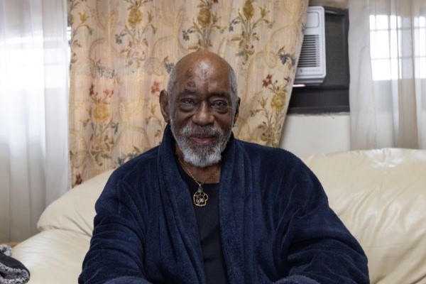 An older black man dressed in a bathrobe sits on a couch, smiling at the camera.