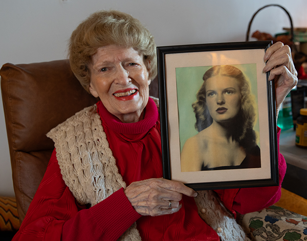 Carol, a senior in NYC. She has light skin, blonde hair and red lipstick. She wears a red turtleneck and white vest. She's holding up a photo of herself as a glamarous young woman.