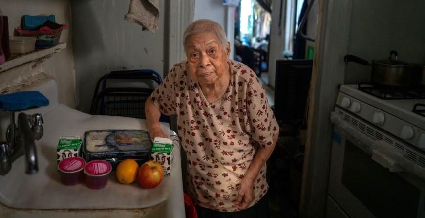 An older Asian American woman standing in her kitchen, a home-delivered meal next to her on the counter.