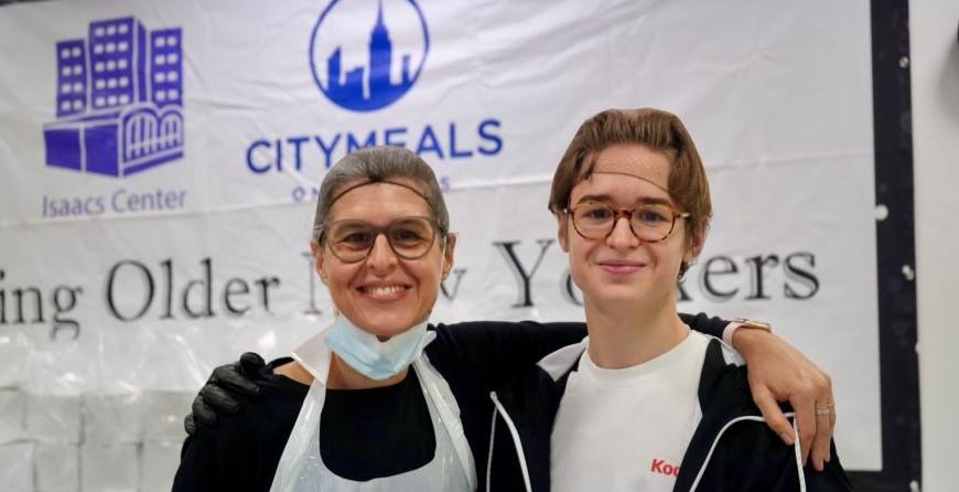 Two kitchen volunteers, dressed in aprons, smile at the camera. 