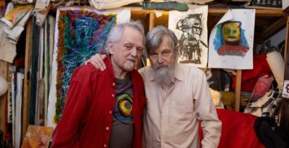 Joel and Jim stand together in their living room, arms around one another. 
